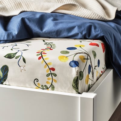 A bed with colourful floral KRONBUSKE fitted sheet in beige, yellow, blue, red, green. Fits thick mattresses up to 26 cm.