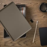 Wooden desk with laptop, notebook, pencil, phone, and box lid. Laptop open, items neatly arranged.
