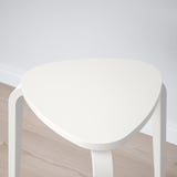A stackable, three-legged stool with a triangular seat is displayed against a wall. The modern design ensures stability and functionality.
