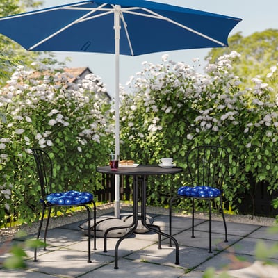 Cozy patio setup with blue umbrella, round table, and two chairs with patterned cushions surrounded by blooming white flowers.