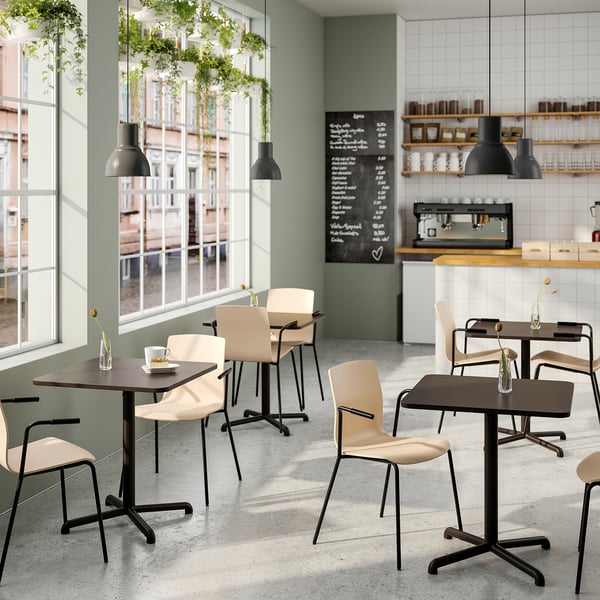 Modern café with wooden tables and beige chairs. Hanging plants, large windows, black lamps, and blackboard menu create a relaxed atmosphere.