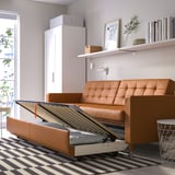 Modern tan leather sofa transforms into bed. Includes storage for bedding. Grey and white striped rug beneath. White cabinet and shelves nearby.