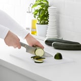 Person slicing zucchini on white board with knife in kitchen.