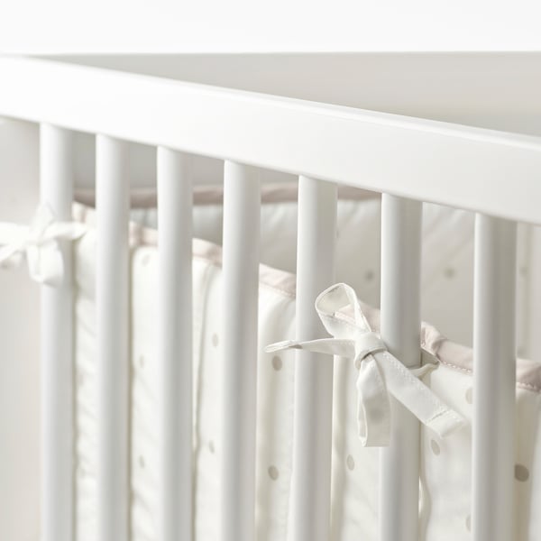 A white crib bumper with ribbons tied to vertical bars, designed to protect a baby from hitting the hard slats.