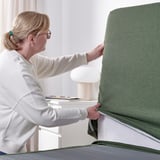 Woman adjusting green fabric divan bed with sloped headboard and multiple cushions.