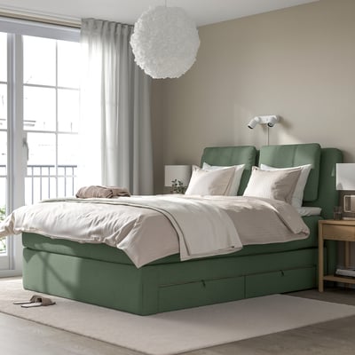 Modern green upholstered bed with sloped headboard, white linens, and integrated storage, positioned near large windows.