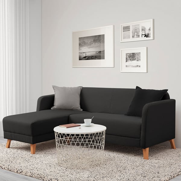 Modern living room: l-shaped black LINANÄS sofa, gray pillows, white round coffee table beneath wall art on light wall. Cozy, compact.