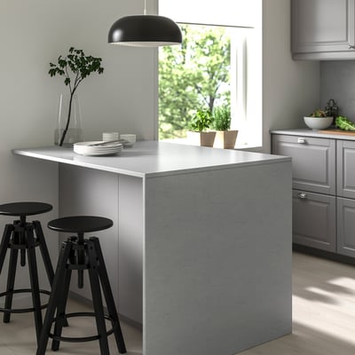 Modern kitchen with large grey LOCKEBO recycled glass island, black barstools, hanging lamp, plates, cups, and potted plants near window.