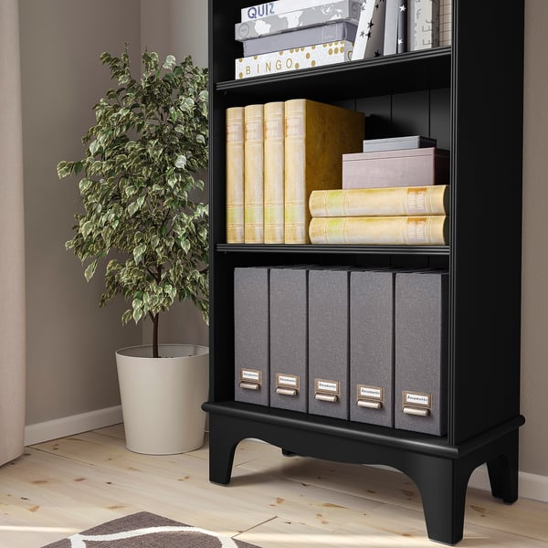 A black bookshelf with three shelves, storing various items like books, decorative boxes, and files. A green potted plant stands beside it.