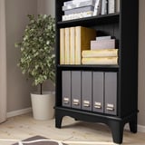 A black bookshelf with three shelves, storing various items like books, decorative boxes, and files. A green potted plant stands beside it.