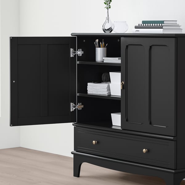 Black cabinet with open doors revealing shelves holding books and pens. Gold handles and a drawer below. The cabinet must be wall-fixed for safety.