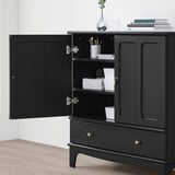 Black cabinet with open doors revealing shelves holding books and pens. Gold handles and a drawer below. The cabinet must be wall-fixed for safety.