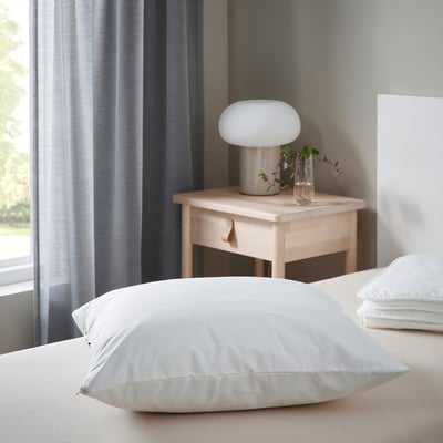 Modern bedroom: white bed with LUDDROS pillow protector, wooden nightstand, lamp, and vase. Protector is large, fits bed perfectly.