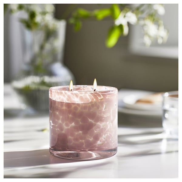 Pink glass candle with floral pattern, two wicks, on white table, surrounded by greenery.
