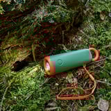 Green flashlight with orange accents and lanyard, resting on mossy forest floor. Quality materials for durability.