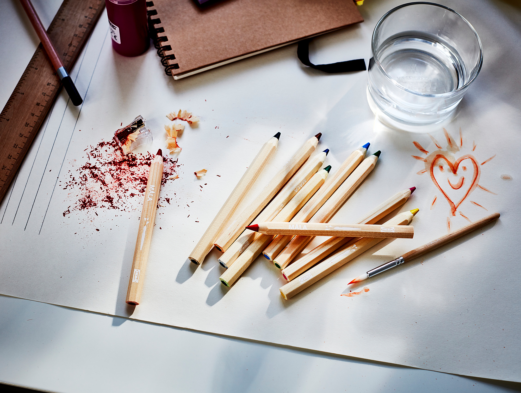 Messy art desk with MÅLA pencils, water glass, sketches, and tools.