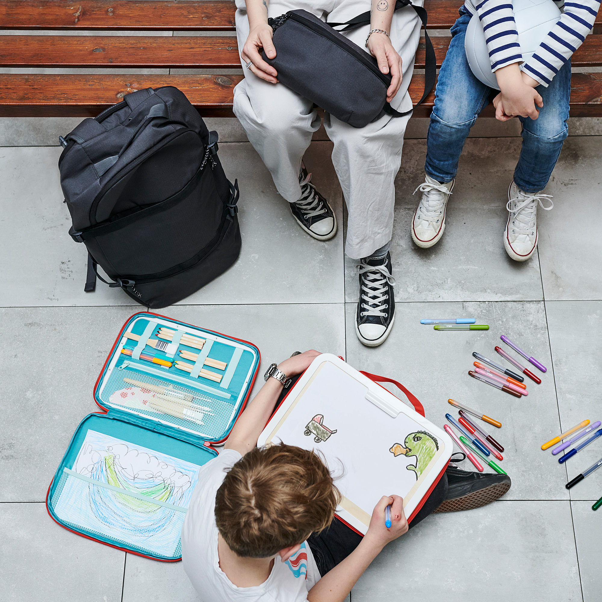 Children on bench, one drawing with open MÅLA art case. Colorful supplies and artwork on ground, large backpack beside them.