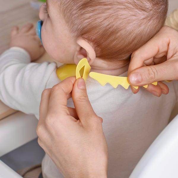 Baby holding a yellow spoon, being fed by an adult wearing a white bib.
