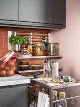 Modern kitchen with grey cabinets, pink walls, and open shelves displaying jars, herbs, fruits, and appliances, featuring led under-cabinet lighting.
