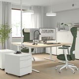Modern home office with MITTZON desk, white with birch veneer. Green ergonomic chair, large dual monitors, and white file cabinet.
