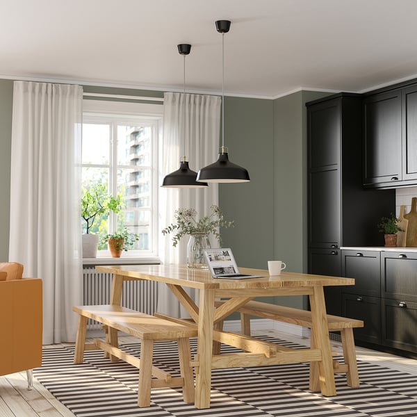 Cozy dining nook with MÖCKELBY table, benches, and pendant lamps. Rug, cabinets, flowers, and a magazine. Brightened by a window with curtains.