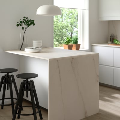 Modern kitchen with large white MÖCKLARP high-pressure porcelain island, black stools, white pendant lamp, green plant, and white cabinets.