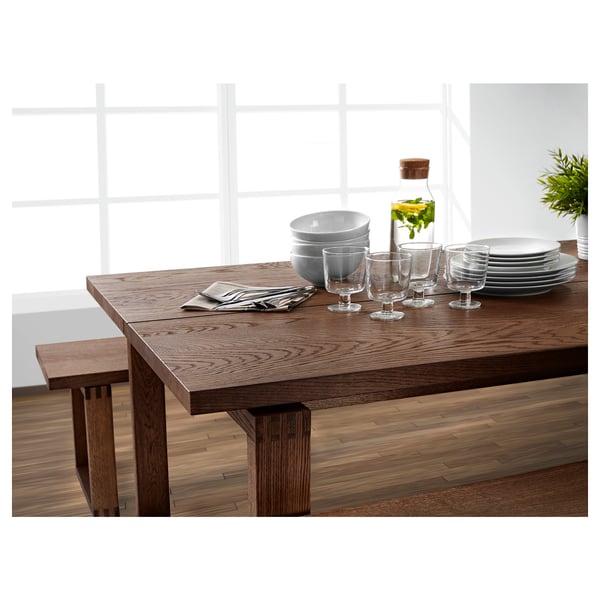 Modern wooden dining table set with clear glasses, white dishes, gray napkins, and a bottle with greens.
