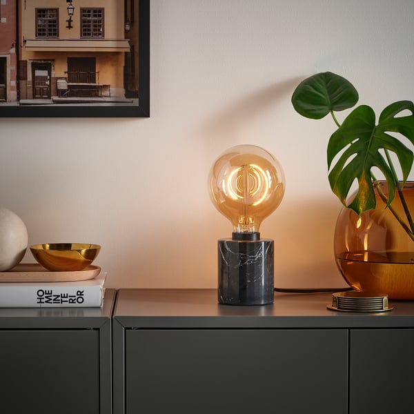 Stylish lamp MOLNART with unique filament, dark console, books, gold bowl, and green plant in warm setup.