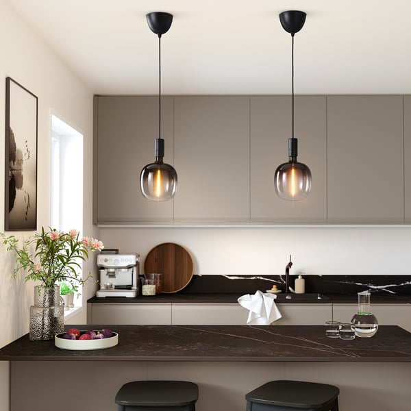 Modern kitchen with black pendant lights, stools, fruits, vase. Illuminated by sleek glass lights, artwork, cabinets in the background.