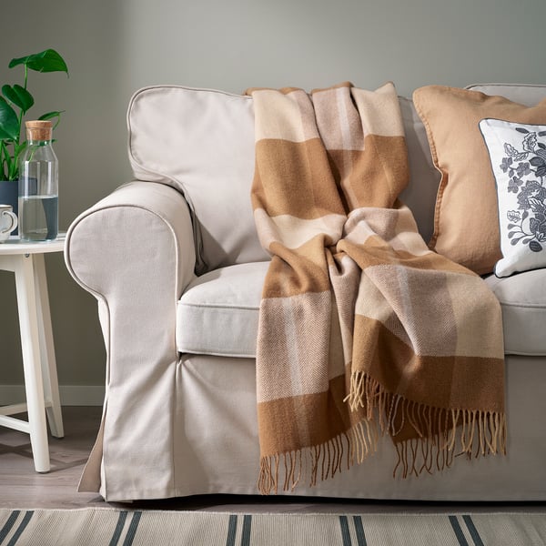 Beige sofa with brown-and-beige checkered wool throw and pillows, white side table, green plant.