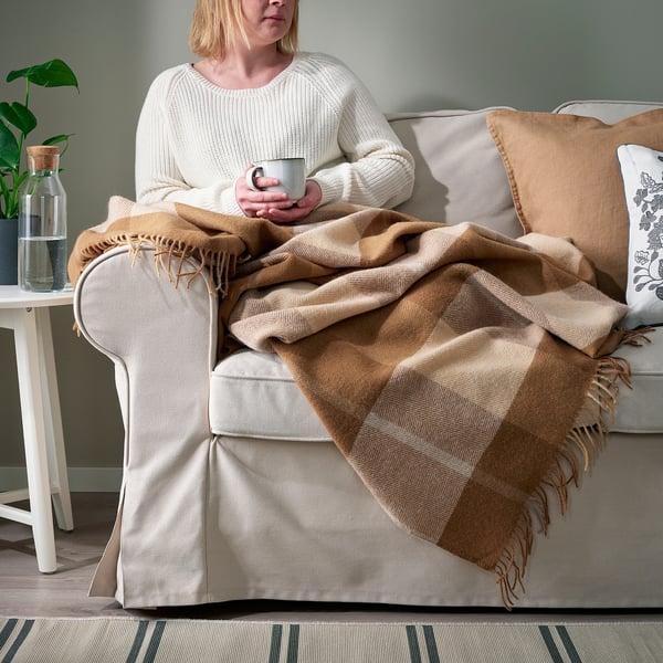 Woman sitting on sofa with warm, light brown MYRULL wool throw blanket, holding a mug, enjoying cozy relaxation.
