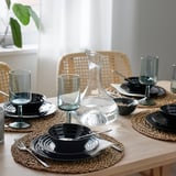 Table set with black glass dinnerware, stackable and dishwasher safe, on woven placemats with clear glasses and water jug.