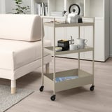 NISSAFORS beige trolley, small, 3-tiered, metal, near white sofa, holding kitchen items.