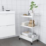 A white NISSAFORS storage trolley with three shelves. Its small but sturdy, holding towels, baskets, and a plant.