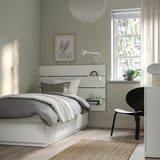 Modern bedroom with white NORDLI bed, headboard, drawers. Black chair by window. Green walls, plant, frame. Bed is focal point.