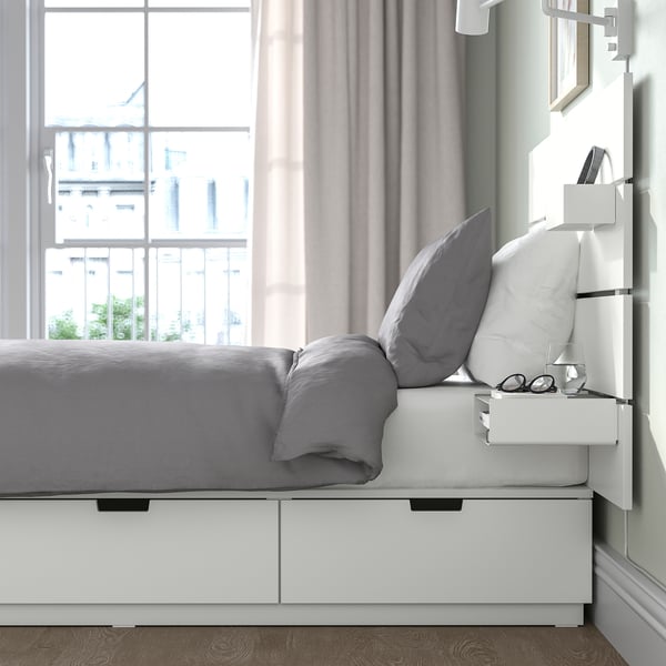 Bedroom scene with white bed frame featuring drawers underneath, a headboard with a box and shelf. Nearby window with curtains.