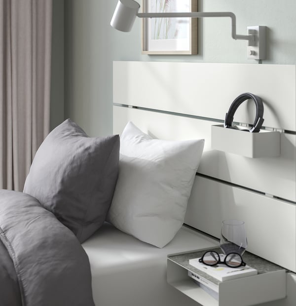 Modern bedroom setup featuring a bed with soft gray and white pillows, a wall-mounted headboard with integrated shelf and reading light.