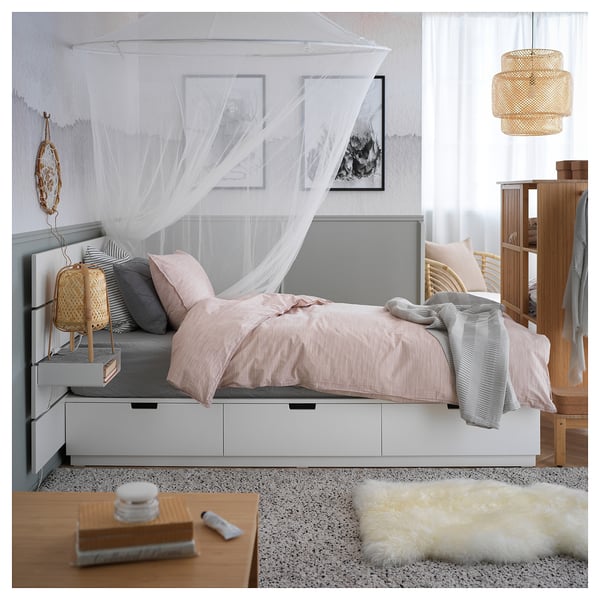 Cozy bedroom with white NORDLI bed, light pink linens, sheer canopy, decorative pillow, wooden side table, lamp, rug, hanging decoration, and shelf.