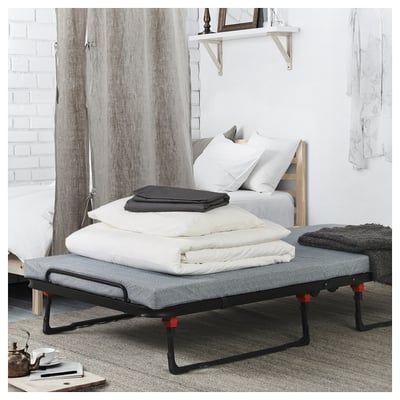 Modern bedroom with white bed, grey fabric guest bed, red feet, folded blankets on top. White curtains, wooden shelf with mirror.