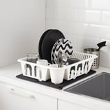 White dish rack on kitchen counter holds cups, pans, and utensils. Black drying mat underneath catches drips.