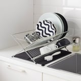 Metal dish rack on a kitchen counter with plates and cups drying; black mat underneath to absorb water.