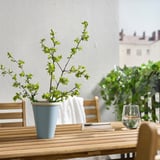 Wooden table holds blue vase with green plant. Next to a wine glass and notepad. Behind, chairs and building.