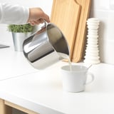 Person pouring liquid from shiny metal pitcher into white mug on counter.