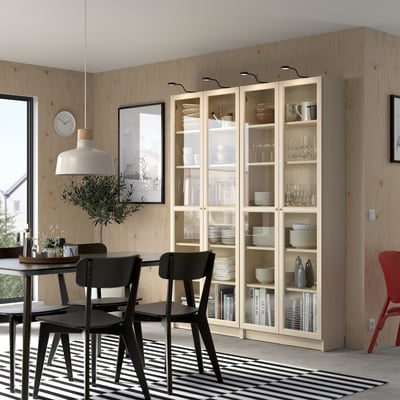 Modern dining area with OXBERG bookcase, wooden table, black chairs, and striped rug. Potted plant and wall clock enhance decor.