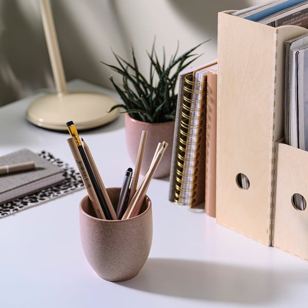 A desk setup with a pink PÄRLHÖNA candle, holding pens, next to a plant and binder.