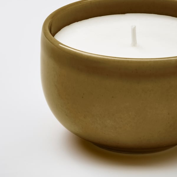 A round stoneware candle holder with a speckled green glaze, containing a white scented candle with a wick.