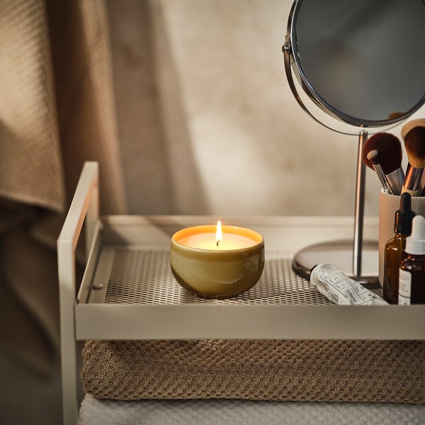 Small lit candle in yellow stoneware bowl on white tray with makeup items.
