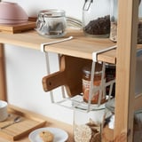 Shelf organizer clips on basket for space optimization, holding jars and wooden utensils beneath shelf.