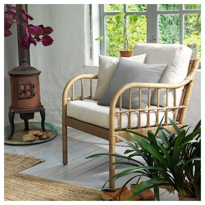 Cosy armchair with light cushions, wicker design, wooden floor; indoor plants, white walls.