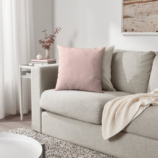 Cozy living room with gray sofa, pink/beige cushions, cream blanket, side table with vase and books.